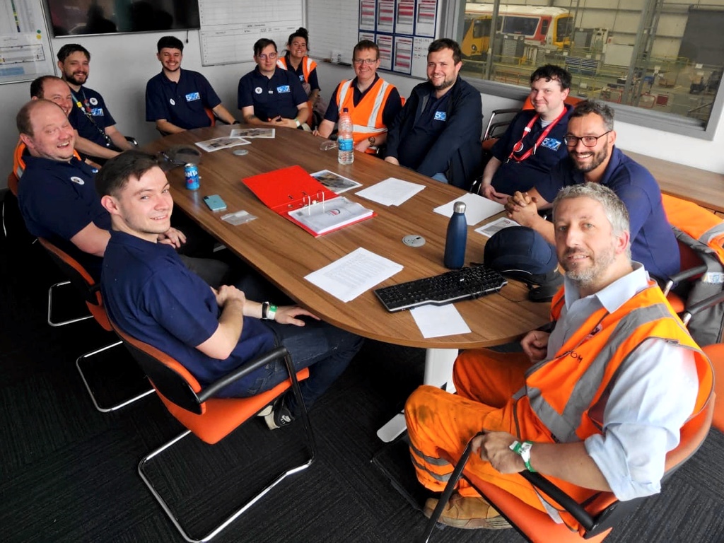 Photo of the C507PS team sitting around a conference table in an office, with the 507 and 465 visible out of the window in V Shop beyond  